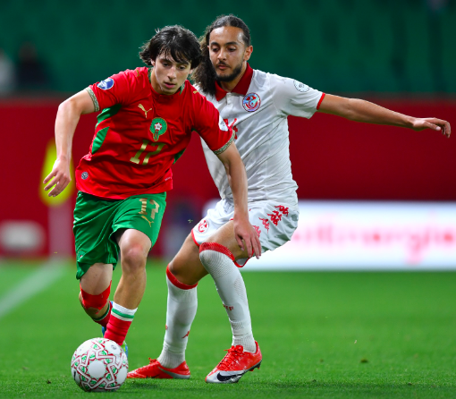 Two soccer players chase the ball during a match; one in red‑green kit, the other in white, on a green field.