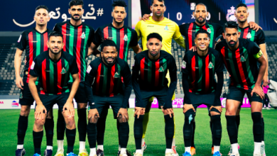 Eleven soccer players in black jerseys with green and red stripes pose for a team photo on a soccer field, with the goalkeeper in yellow in the back row.