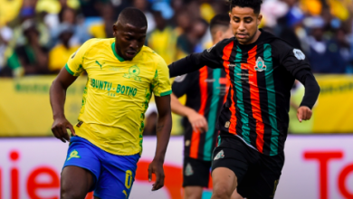 Soccer players sprint for the ball during a match; left in yellow jersey with blue shorts, right in black with red-and-green stripes; blurred crowd in stands behind.