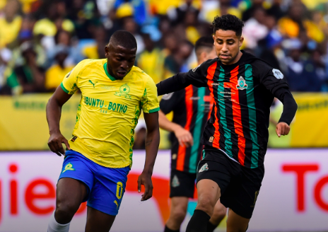 Soccer players sprint for the ball during a match; left in yellow jersey with blue shorts, right in black with red-and-green stripes; blurred crowd in stands behind.