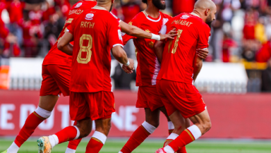 Five soccer players in red jerseys celebrate a goal on a green pitch with a cheering crowd in the stands.