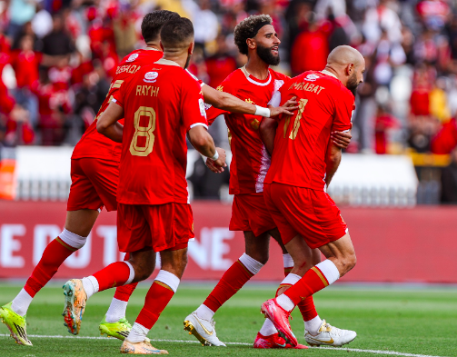 Five soccer players in red jerseys celebrate a goal on a green pitch with a cheering crowd in the stands.