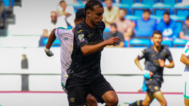 Soccer player in a black kit dribbles the ball during a match, with teammates and blue stadium seats in the background.