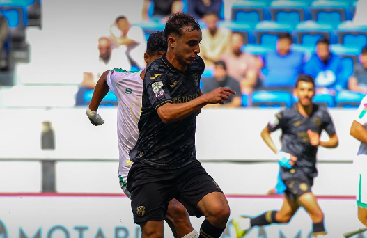 Soccer player in a black kit dribbles the ball during a match, with teammates and blue stadium seats in the background.