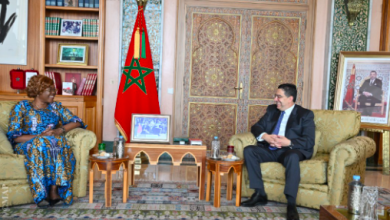 | MCG24 | موقع إلكتروني مغربي شامل متجدد على مدار الساعة Two people in a formal room: a woman in a blue patterned dress on the left and a man in a suit on the right, seated on beige armchairs, with a Moroccan flag and ornate doors behind them.