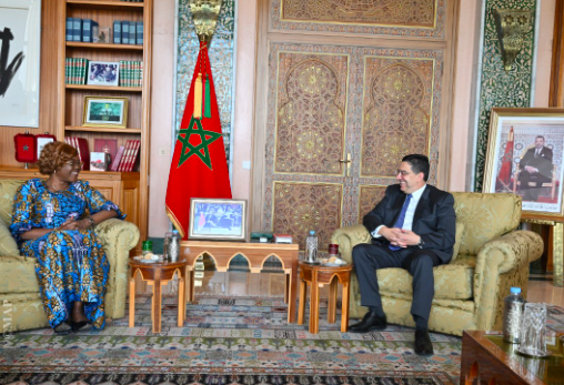 Two people in a formal room: a woman in a blue patterned dress on the left and a man in a suit on the right, seated on beige armchairs, with a Moroccan flag and ornate doors behind them.