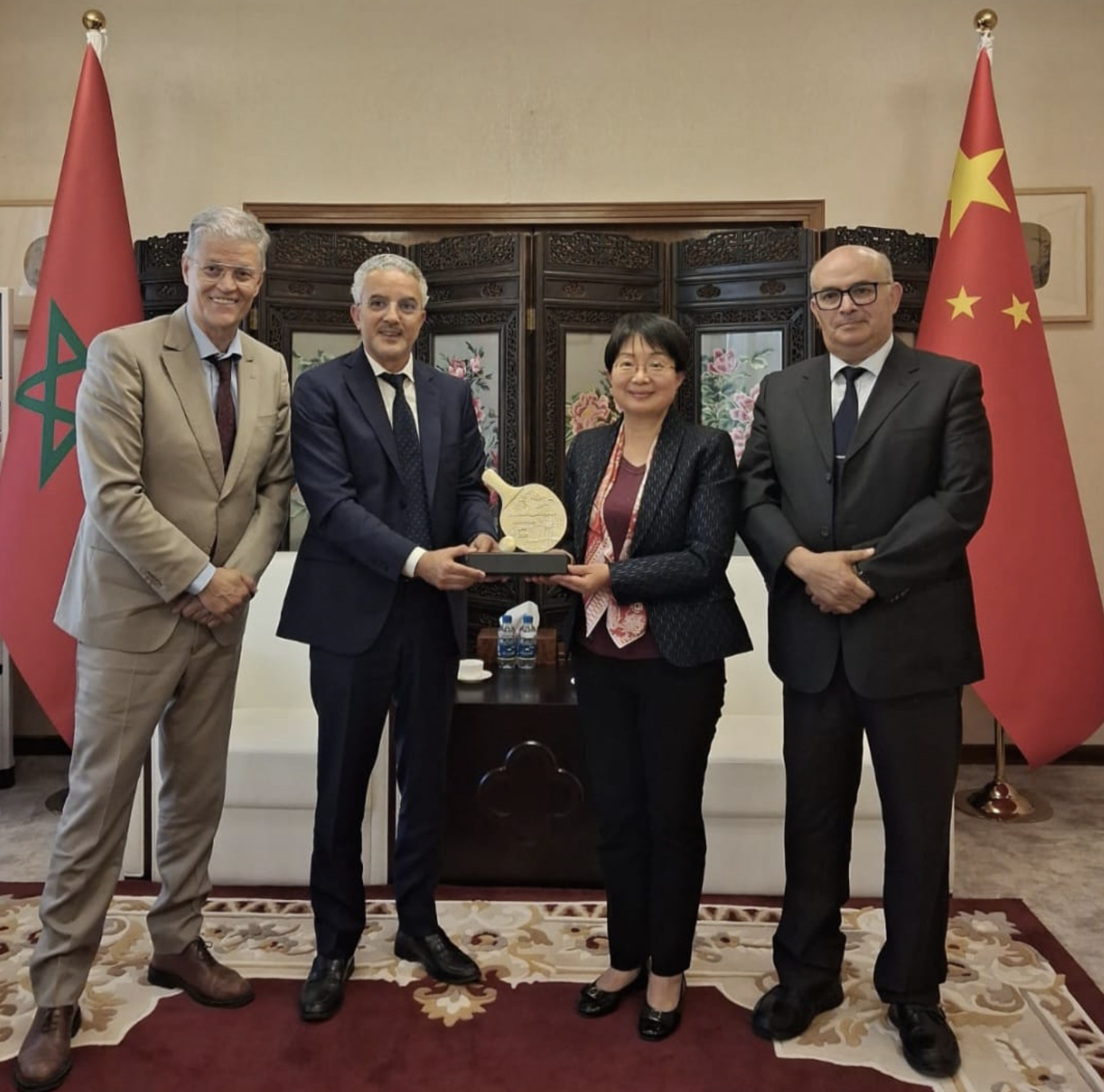 Four professionals pose indoors with flags (Morocco left, China right) as they display a wooden award plaque together in front of a carved screen.