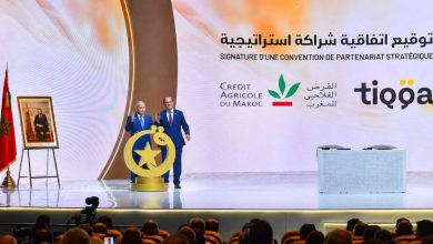 Two men in suits on a bright stage at a formal signing ceremony, with a Moroccan flag, portrait on an easel, and a large backdrop with Arabic text and logos.