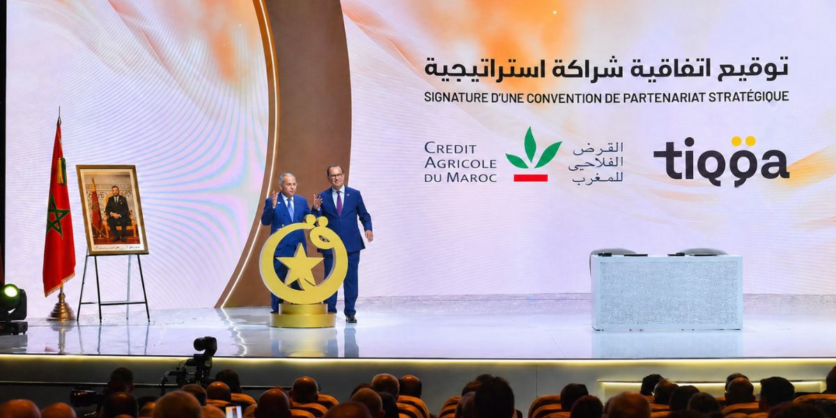 Two men in suits on a bright stage at a formal signing ceremony, with a Moroccan flag, portrait on an easel, and a large backdrop with Arabic text and logos.