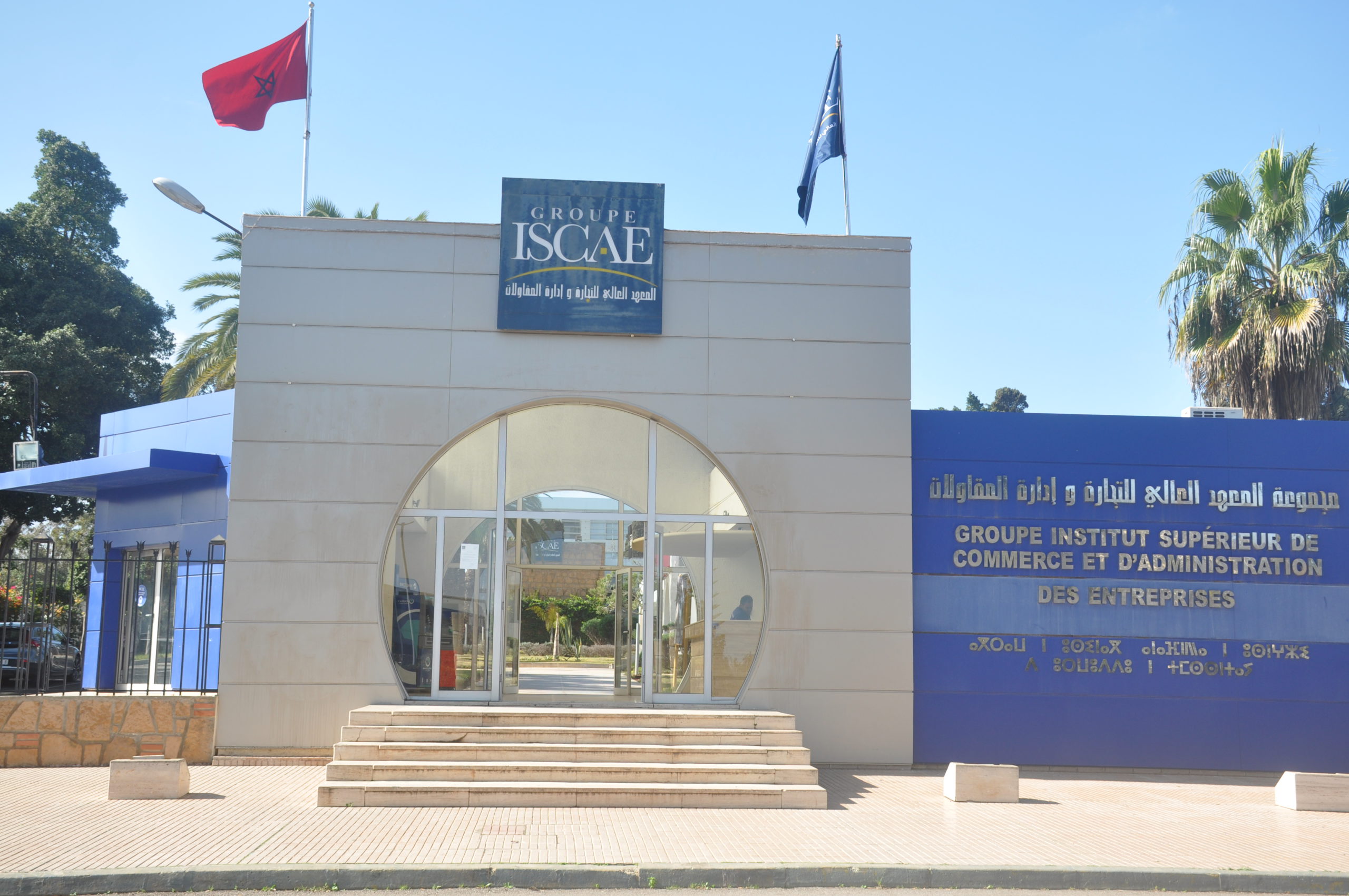 Entrance to Groupe ISCAE building with a blue sign reading 'GROUPE ISCAE' and Arabic text, glass doors, and steps; Moroccan flag on a flagpole to the left.