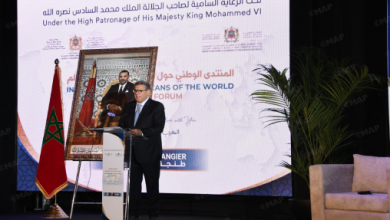 DSC_0819122323 | MCG24 | موقع إلكتروني مغربي شامل متجدد على مدار الساعة Speaker in a dark suit delivering a talk at a podium on a stage, with a Moroccan flag and a large framed portrait behind, and a backdrop referencing a world forum event.