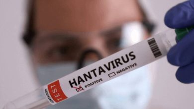 Scientist wearing goggles and a mask holds a test tube labeled HANTAVIRUS with positive/negative checkboxes visible in the foreground.