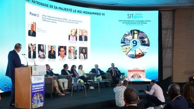 Speaker at a podium addressing a panel of seven seated experts on a stage, with a large screen displaying SITeau conference visuals behind them.