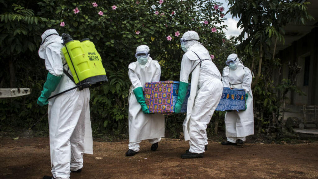 EBOLA-RDC | MCG24 | موقع إلكتروني مغربي شامل متجدد على مدار الساعة Four healthcare workers in full PPE carry a colorfully wrapped coffin along a dirt path.