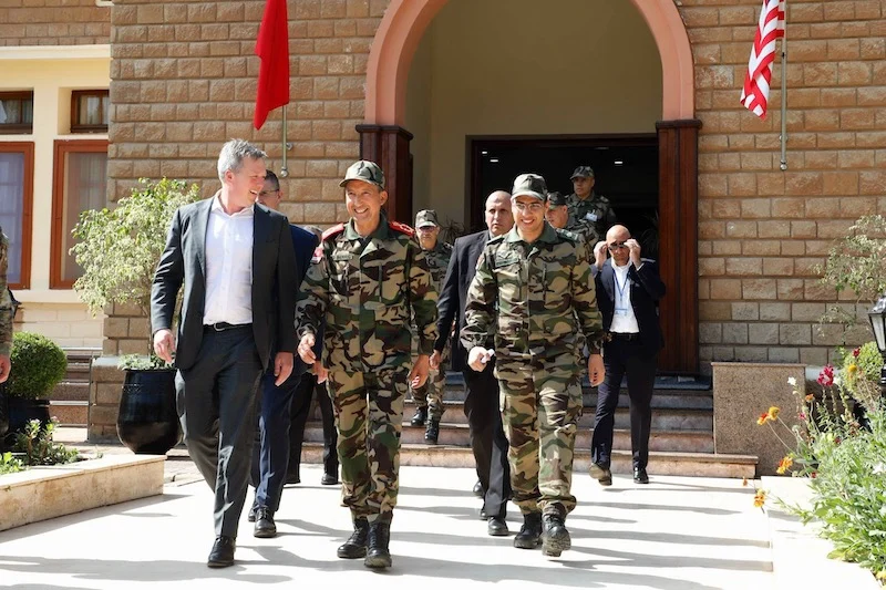 Group of soldiers in camouflage walk with officials down a sunlit entrance steps, flag banners visible on a brick building behind them.
