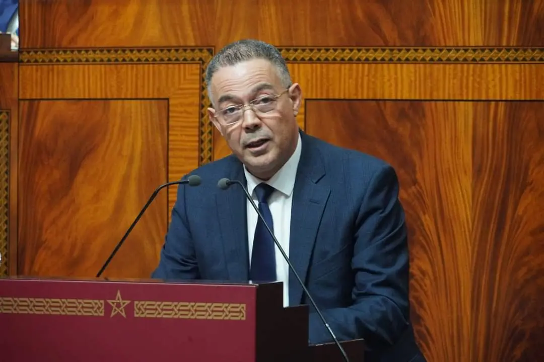FB_IMG_1731690583648 | MCG24 | موقع إلكتروني مغربي شامل متجدد على مدار الساعة Man in a navy suit and glasses speaks behind a wooden podium with two microphones in front of a wood-paneled backdrop.