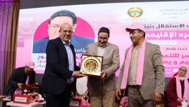 Men in suits present a framed award on stage during a ceremony, with pink banners and a large poster in the background.