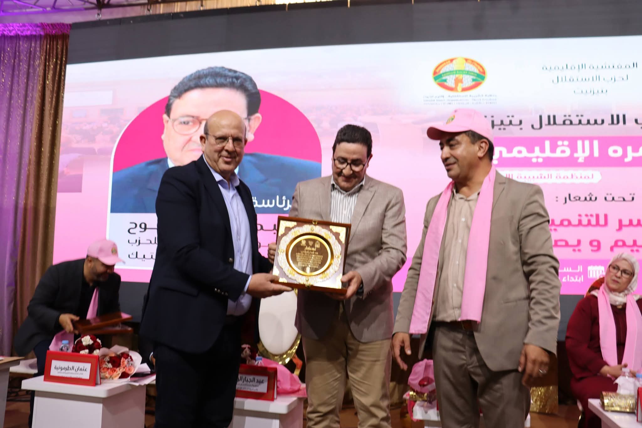 Men in suits present a framed award on stage during a ceremony, with pink banners and a large poster in the background.