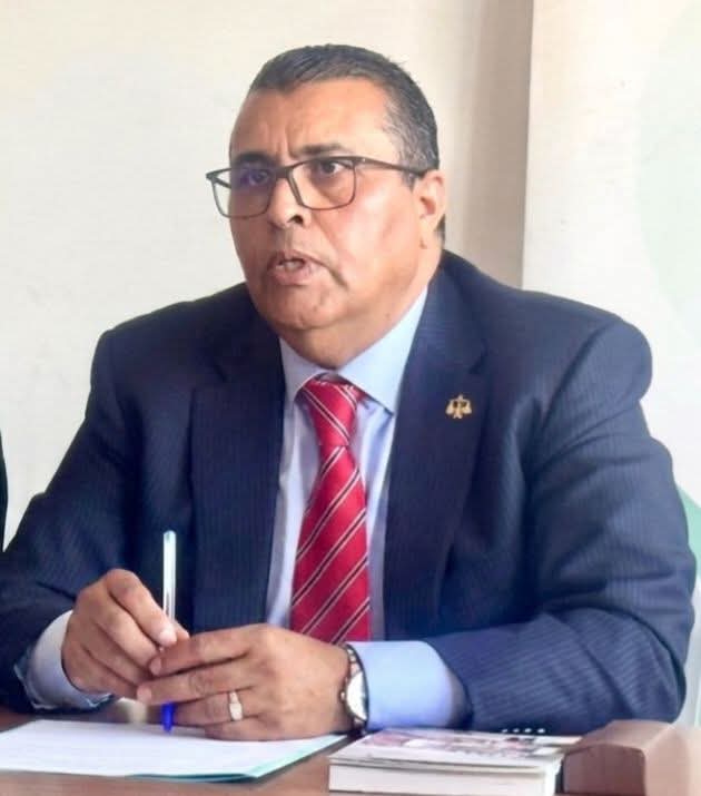 Professional man in a dark suit and red striped tie, wearing glasses, seated at a desk with a pen and papers in front of him on a light background.