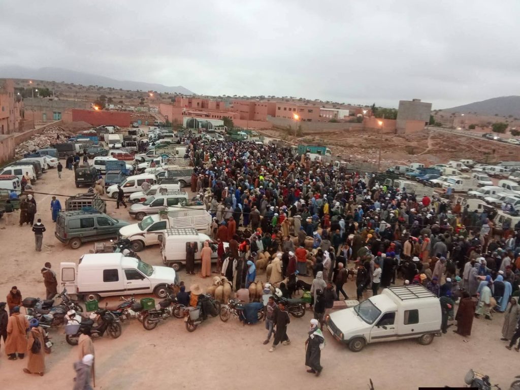 Large crowd of people gathered in a dusty town square, surrounded by cars and vans with orange-brown buildings in the background.