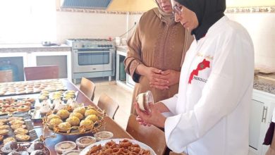 Two women wearing hijabs stand in a bright kitchen, inspecting trays of Moroccan pastries laid out on a table with jars of sweets nearby