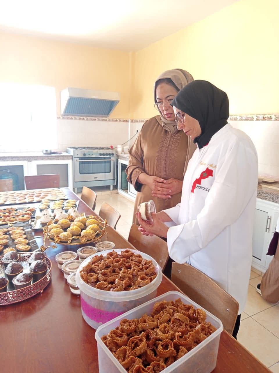 Two women wearing hijabs stand in a bright kitchen, inspecting trays of Moroccan pastries laid out on a table with jars of sweets nearby