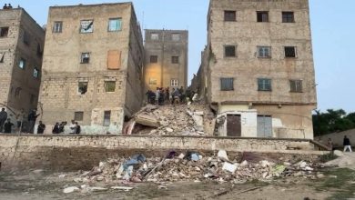 Group of people standing on a large pile of rubble between damaged apartment buildings after a collapse, likely a rescue scene.
