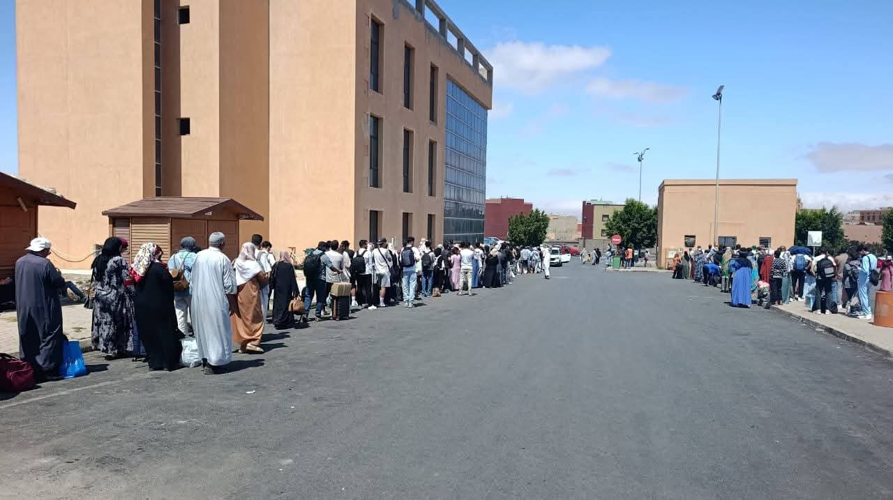 Long queue of people standing along a street outside a large tan building on a sunny day, many wearing traditional robes and head coverings.