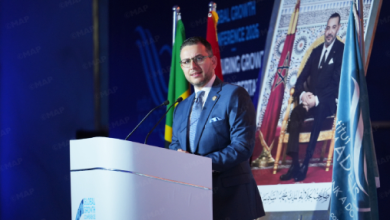 Man in a blue suit giving a speech at a podium during a conference, with national flags behind him and a large portrait on a screen in the background.