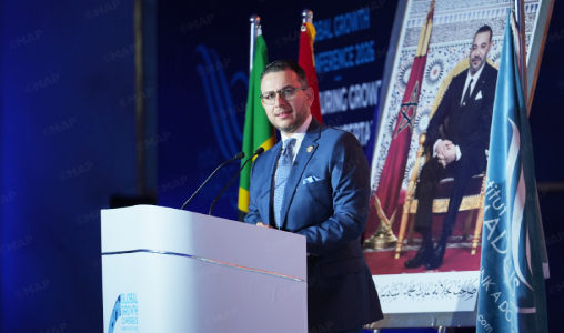 Man in a blue suit giving a speech at a podium during a conference, with national flags behind him and a large portrait on a screen in the background.