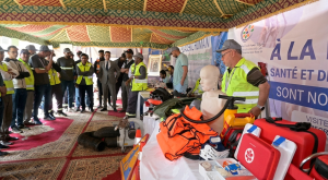 Group of people in high-visibility vests watching an emergency medical equipment display under a decorated tent with a mannequin and first-aid kits on the table.