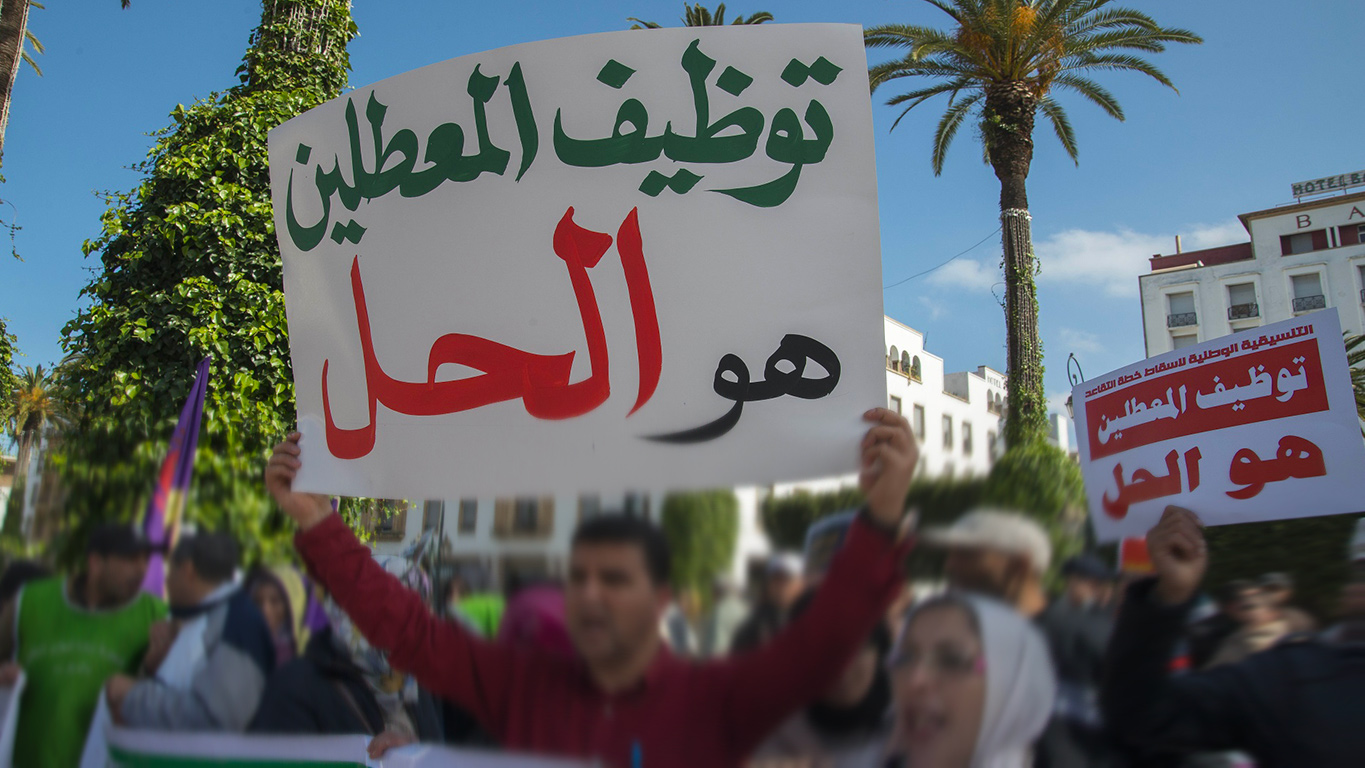 Protest on a sunny street with people marching, holding a large white sign with green and red Arabic text, and a smaller red-and-white sign nearby; palm trees and buildings in the background.