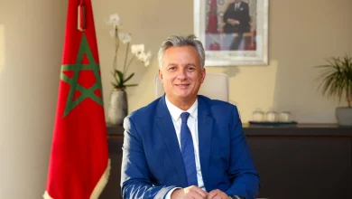 Man in a blue suit sits at a desk in an office with a Moroccan flag and a framed portrait visible in the background.