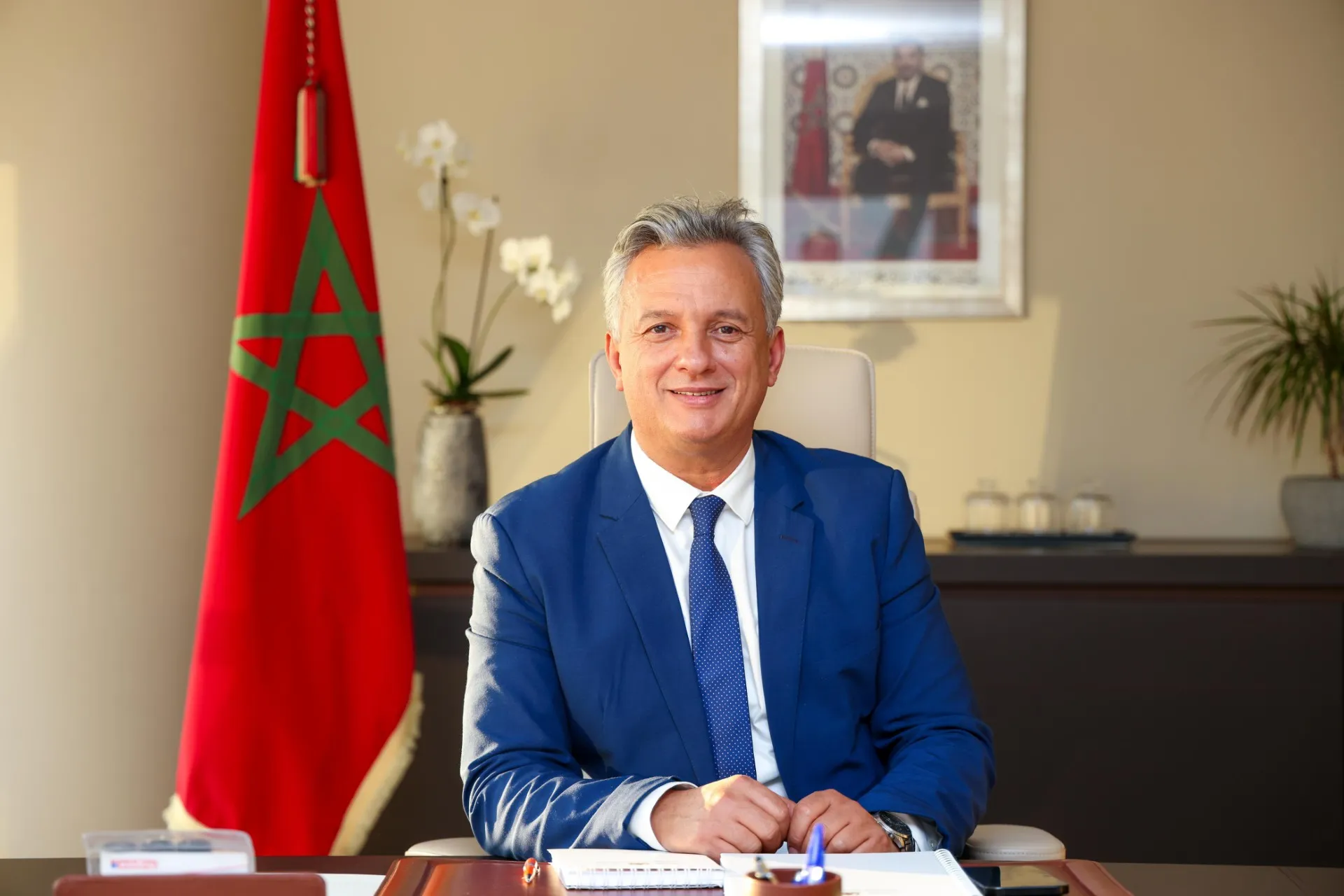 Man in a blue suit sits at a desk in an office with a Moroccan flag and a framed portrait visible in the background.