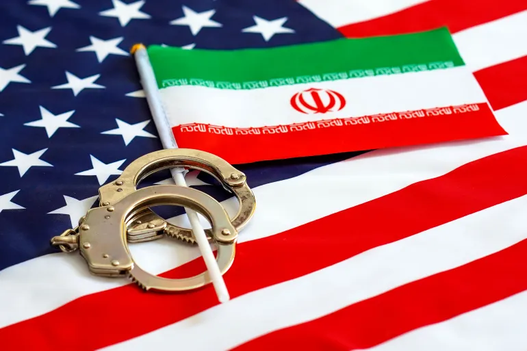 Handcuffs resting on a U.S. flag with a small Iranian flag laid across it, suggesting tension between the two nations.