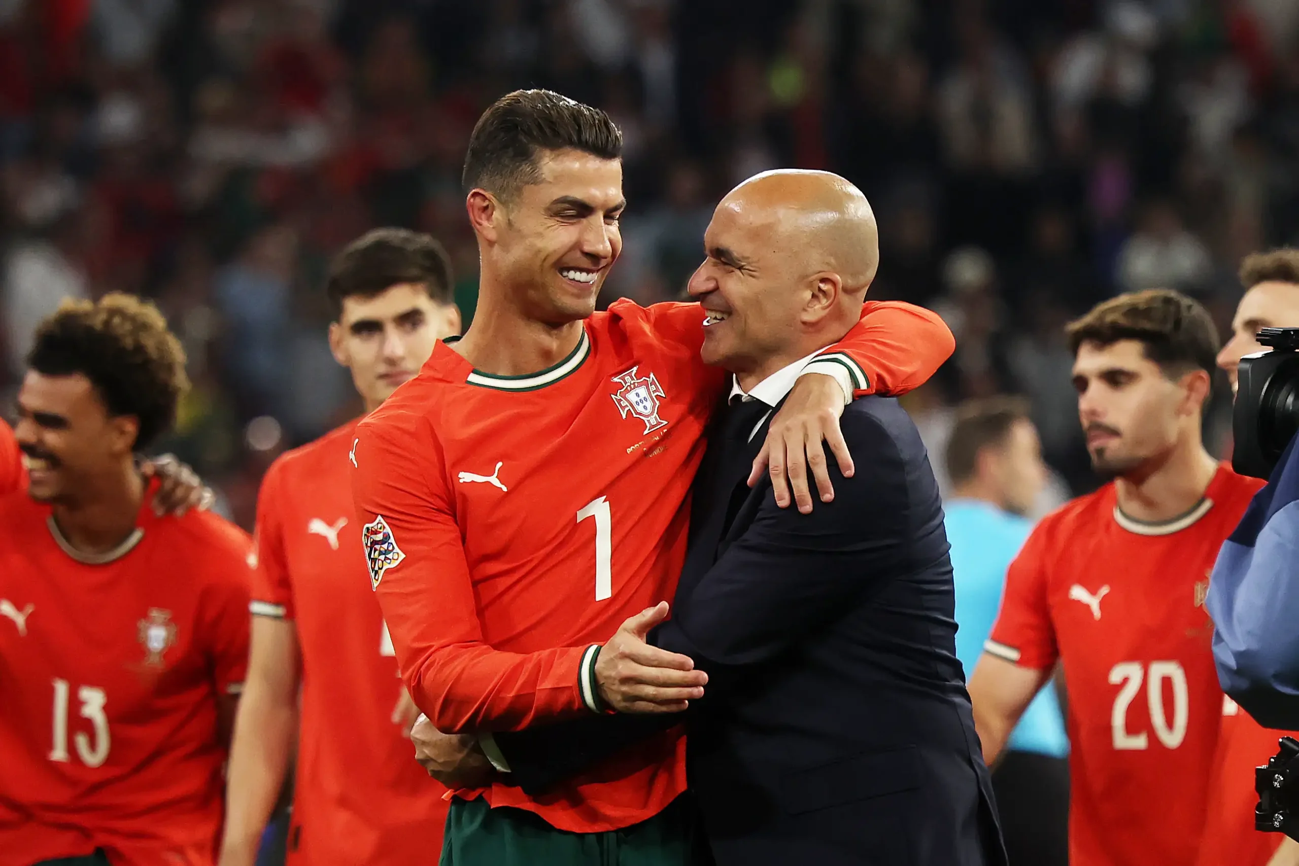 Portugal national team players celebrate on the field, embracing a teammate in red jerseys with numbers visible (7 and 20).