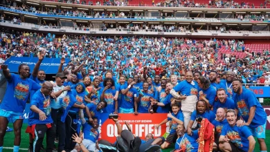 Soccer team and staff celebrate on the field after qualifying, wearing matching blue shirts with a large red 'QUALIFIED' banner and a cheering crowd in the stands behind them.