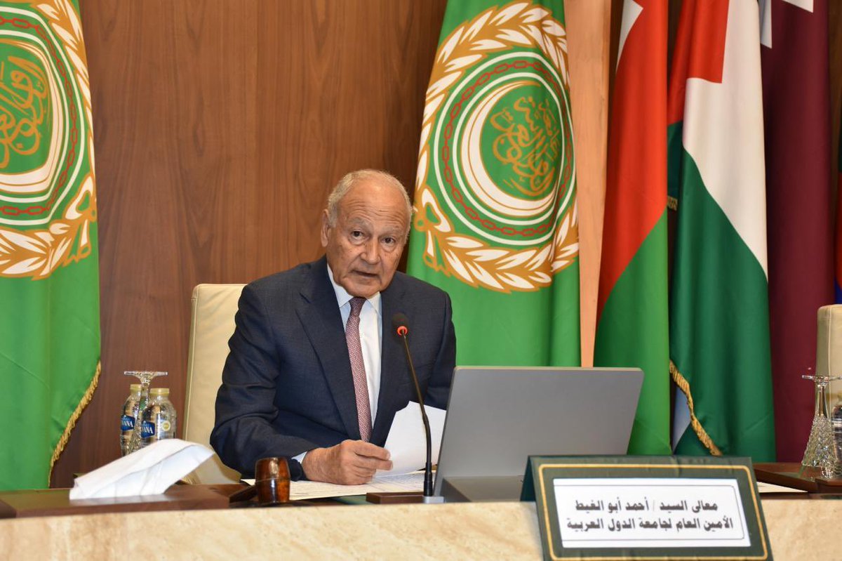 An older man in a dark suit speaks into a microphone at a conference table, with a laptop, papers, and a nameplate in Arabic in front of him, flanked by green flags.