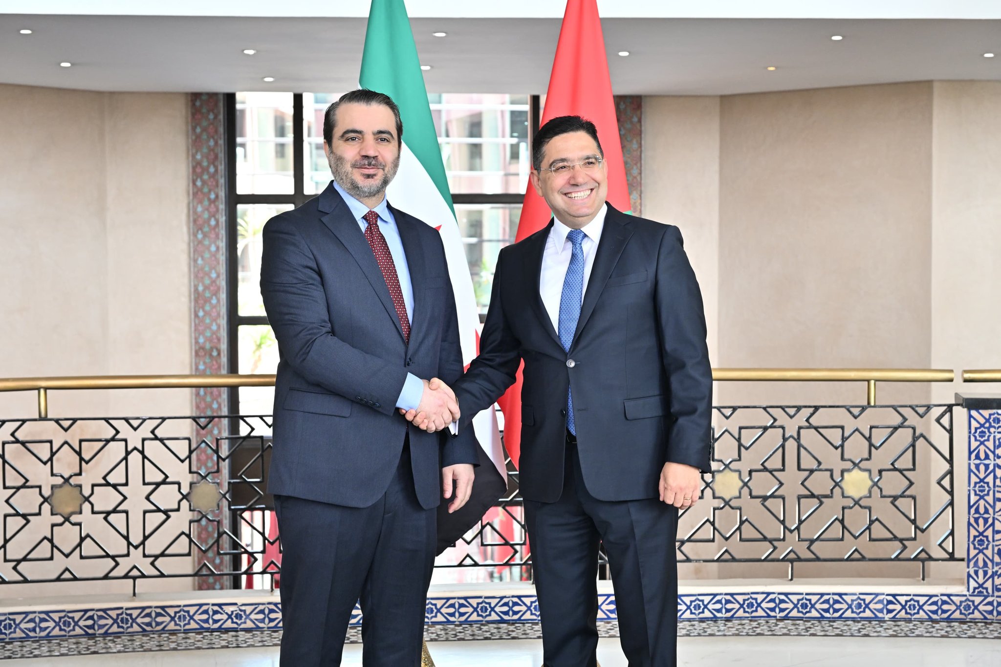 HISGFWLXcAAOsnJ | MCG24 | موقع إلكتروني مغربي شامل متجدد على مدار الساعة Two men in dark suits shake hands in a formal setting with flags behind them.