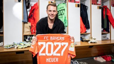 Man in a locker room displaying an orange FC Bayern jersey with 2027 and Neuer printed on it.