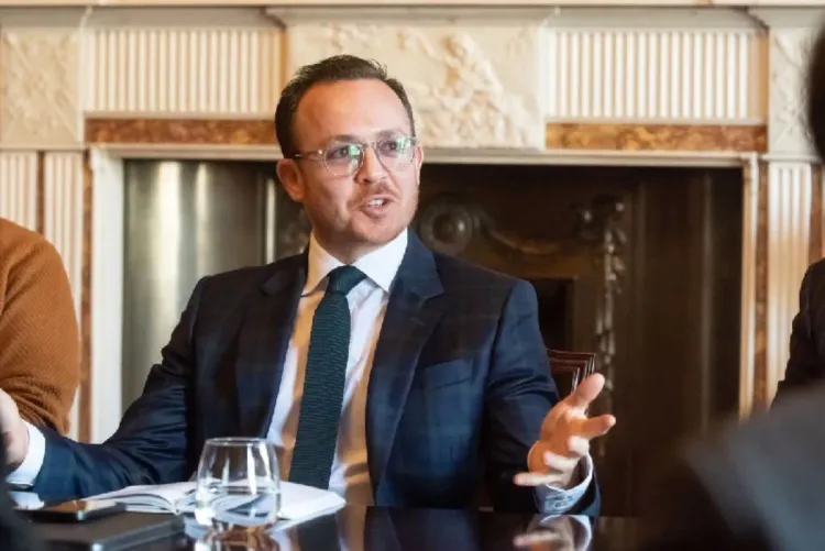 Man in a dark blue suit and teal tie speaks at a formal meeting, gesturing with his hands in front of a fireplace backdrop.