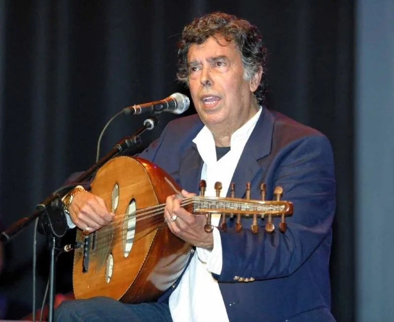Man in a blue blazer and white shirt playing an oud and singing into a microphone on stage