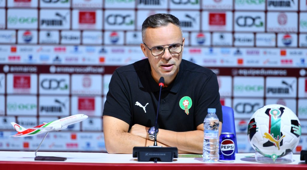 IMG-20260317-WA0024-1024x568 | MCG24 | موقع إلكتروني مغربي شامل متجدد على مدار الساعة Male football official with Morocco crest speaks at a press conference, microphone in front, plus a water bottle, Pepsi can, model airplane, and a football on the table.