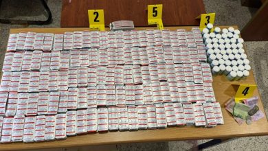 Table loaded with hundreds of small pill bottles laid out in rows, with yellow evidence markers labeled 2, 3, 4, 5 visible in the background and some cash on the side.",
