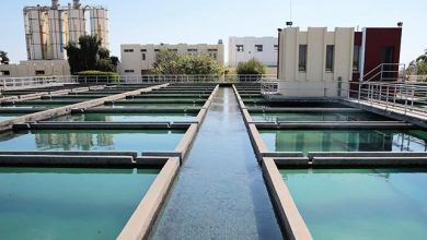 Water treatment plant with parallel rectangular basins and a central channel of flowing water, buildings in the background.