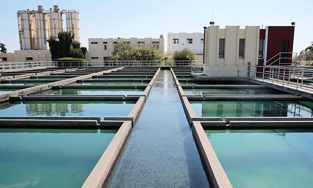 Water treatment plant with parallel rectangular basins and a central channel of flowing water, buildings in the background.