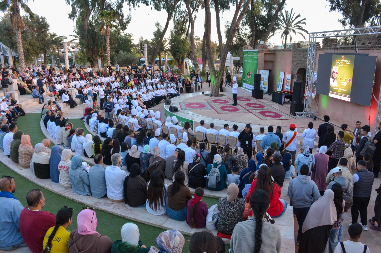 Large outdoor audience seated in a stone amphitheater watching a speaker on stage with a large screen and banners nearby on a sunny day