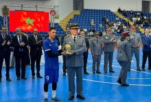 IMG-20260513-WA0366 | MCG24 | موقع إلكتروني مغربي شامل متجدد على مدار الساعة A young athlete in a blue uniform receives a trophy from a uniformed official at a sports ceremony inside a gym, with others clapping and seated spectators in blue seats behind them.