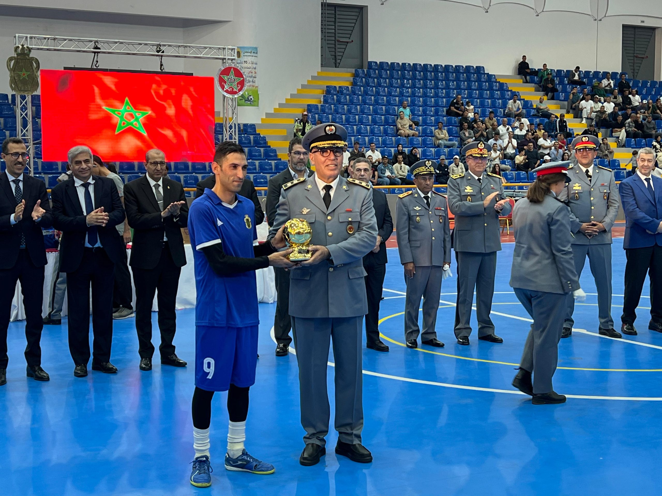 IMG-20260513-WA0366 | MCG24 | موقع إلكتروني مغربي شامل متجدد على مدار الساعة A young athlete in a blue uniform receives a trophy from a uniformed official at a sports ceremony inside a gym, with others clapping and seated spectators in blue seats behind them.