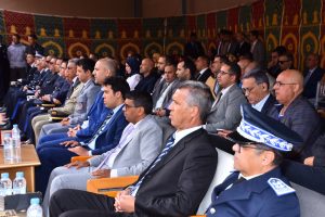 Audience at a formal ceremony: officials in suits and uniforms seated in rows under a colorful patterned backdrop, listening attentively.
